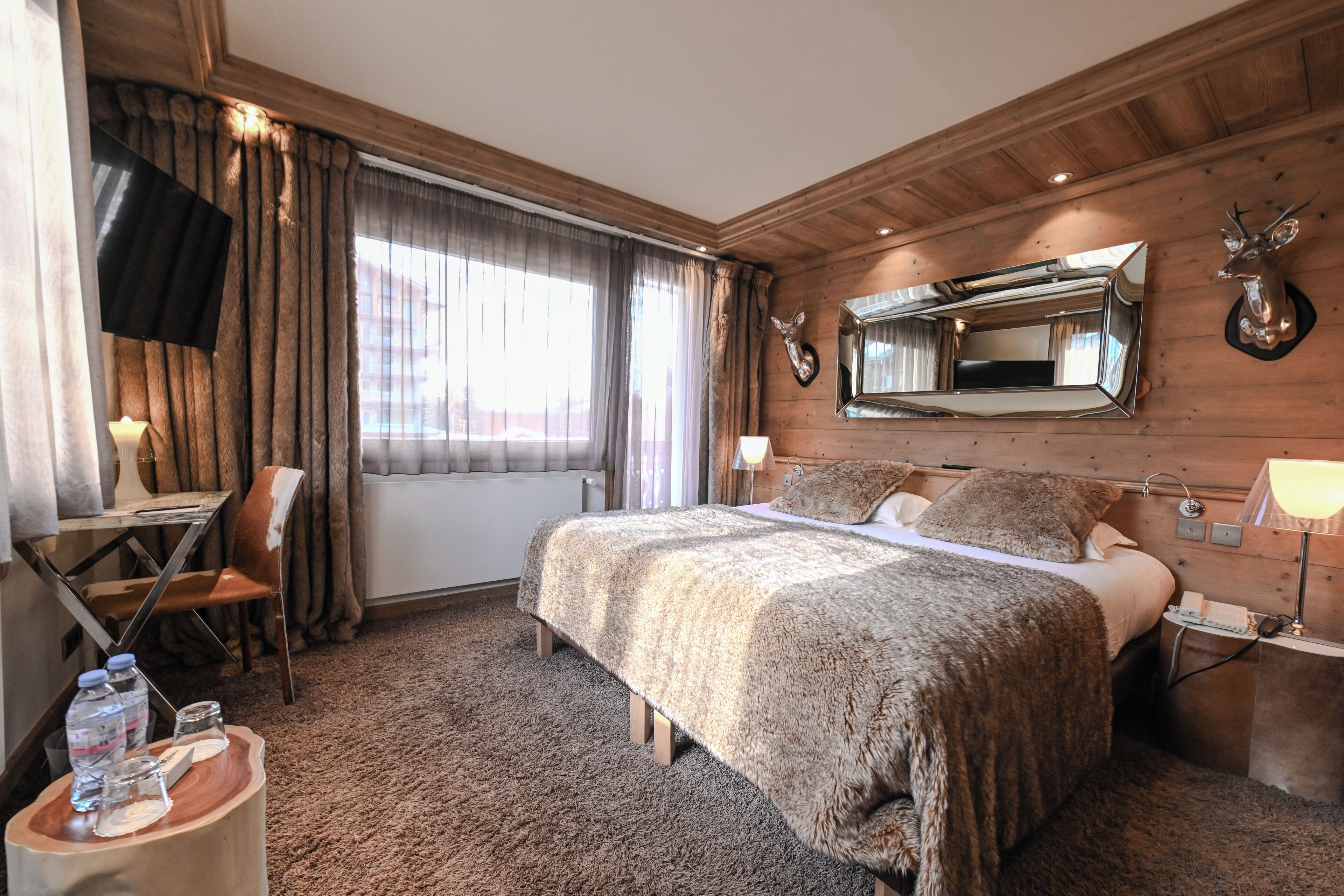 A hotel room with a large bed, a desk with a chair, a TV mounted on the wall, and a mounted deer head.