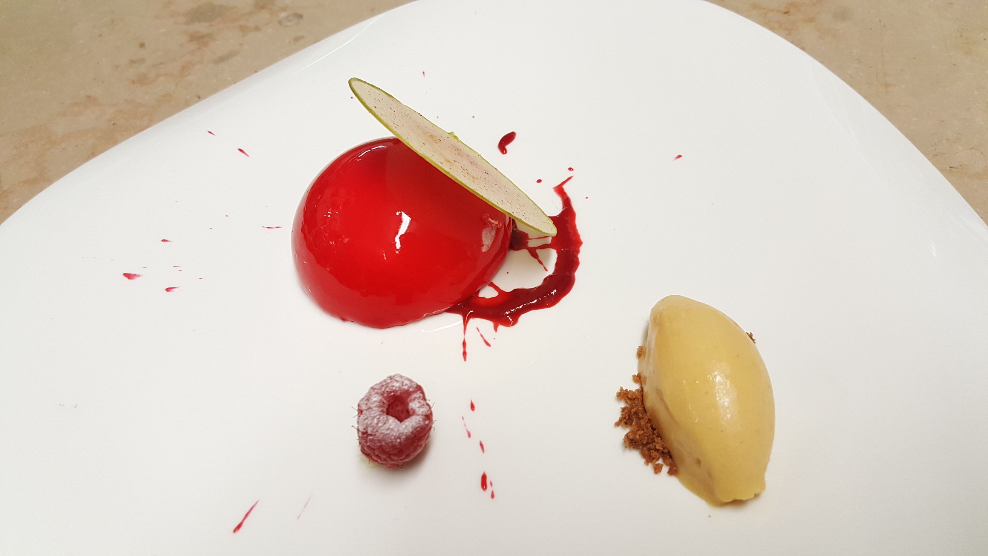 Un plato de postre con una esfera de gelatina roja, una porción de pastel y una frambuesa.