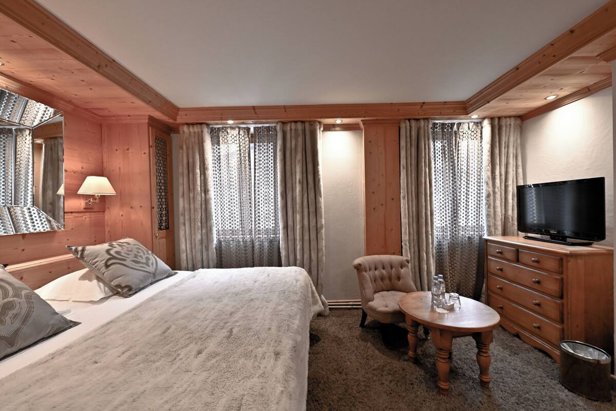 Een hotelkamer met een bed, een televisie, een houten dressoir, een stoel en een klein tafeltje met een lamp.