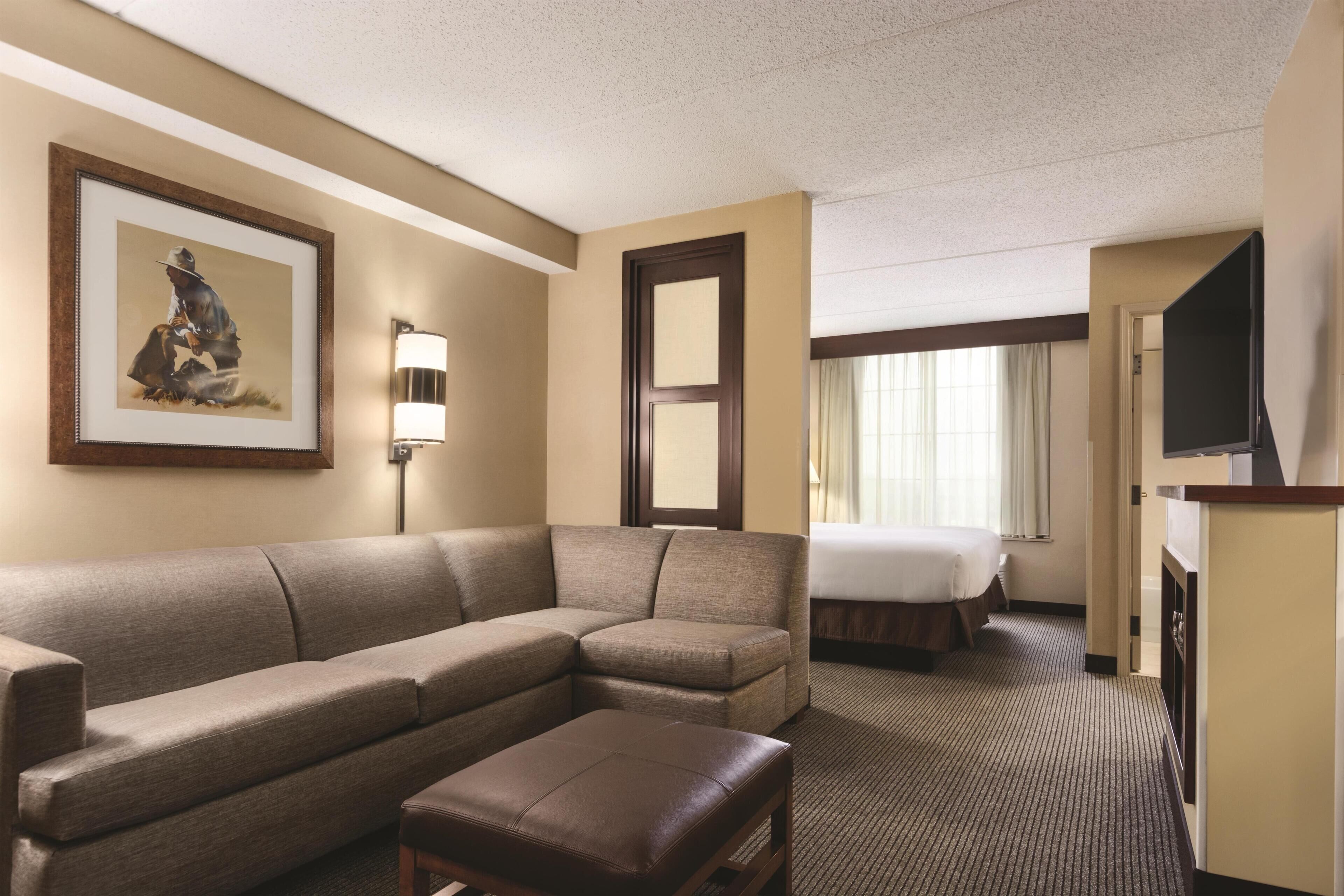 Habitación de hotel con sofá modular, un pequeño reposapiés, cama, televisor y un cuadro en la pared.