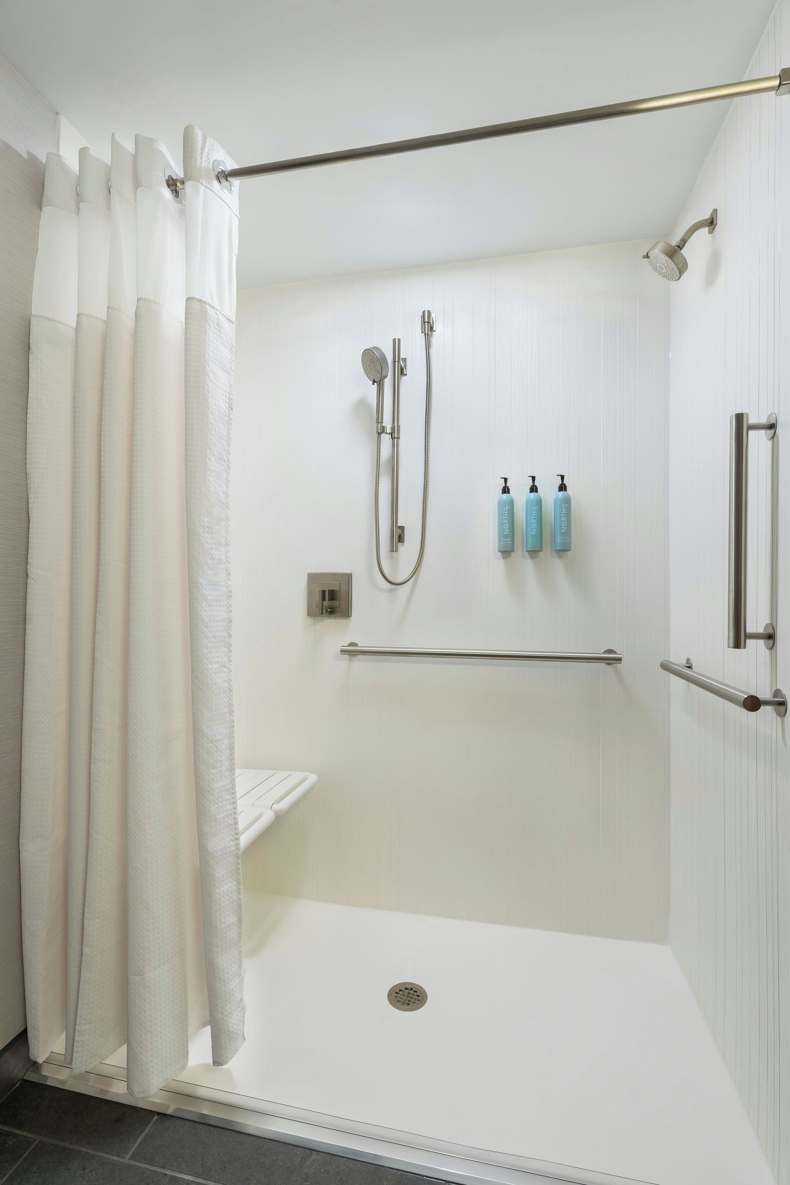 Eine begehbare Dusche mit Glastür, einer Handbrause, einem am Wand montierten Wasserhahn und einem Ablagebrett mit drei Flaschen.