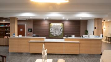 A hotel reception area with a wooden reception desk, a large decorative wall piece, and a seating area with a green chair.