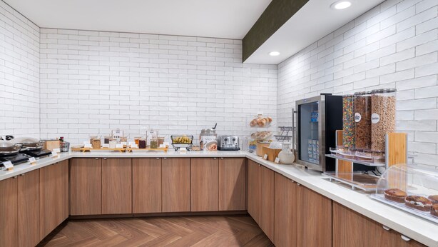 A modern kitchen with white brick walls, wooden cabinets, and a variety of food items displayed on the counter. - Fairfield Inn and Suites by Marriott Denton (Denton)