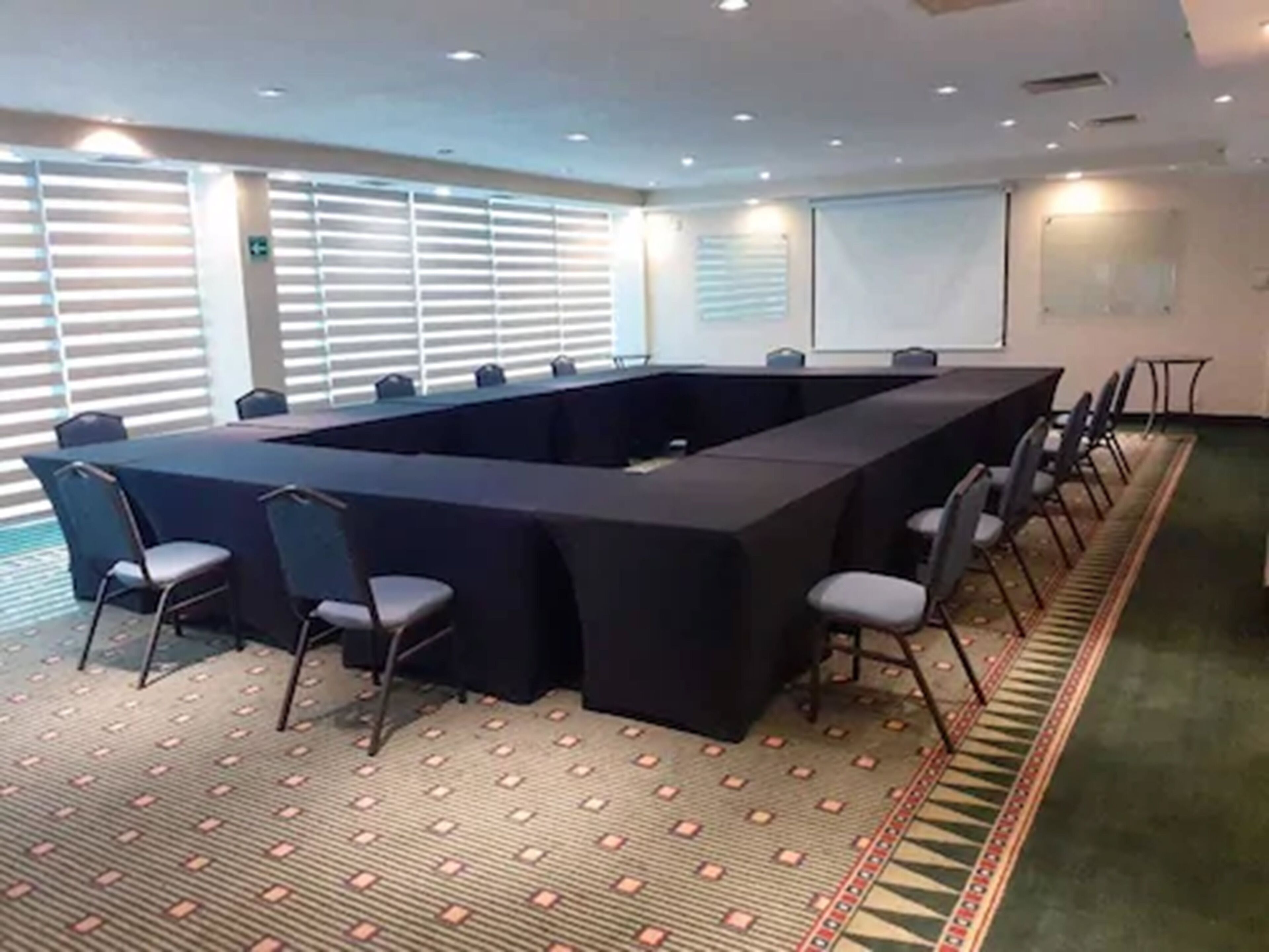 A conference room with a long table and chairs, large windows with blinds, and a patterned carpet.