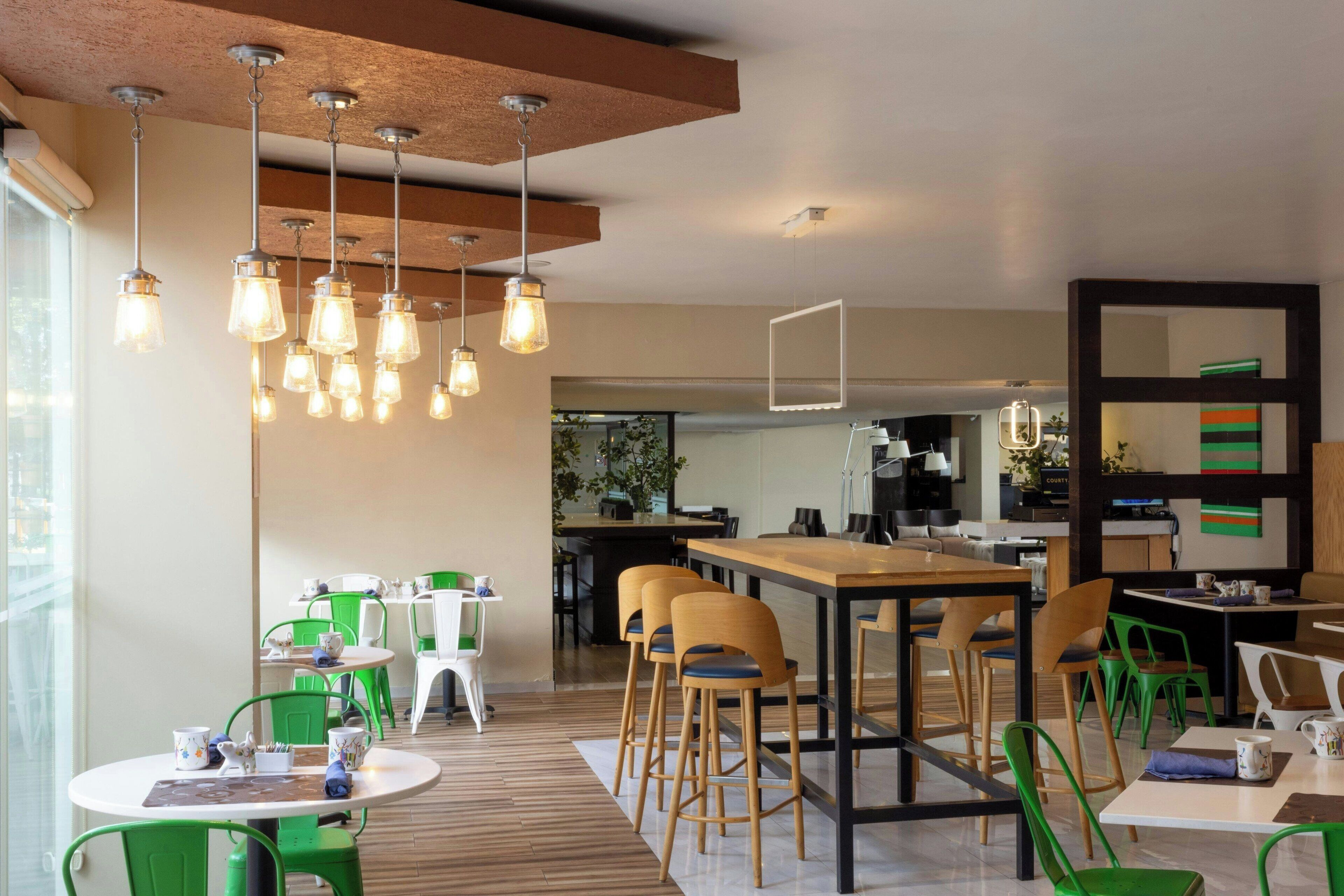 A modern cafe with wooden tables and green chairs, hanging light fixtures, and a view of the kitchen area.