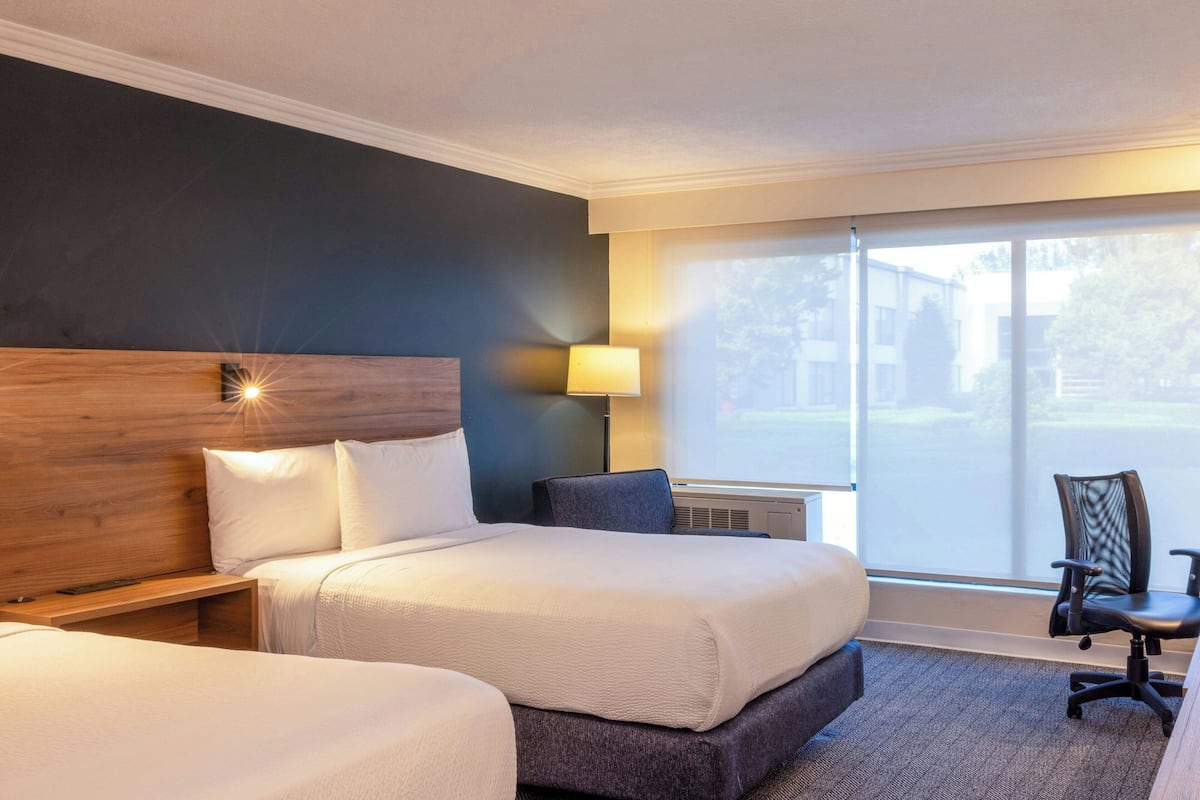 Une chambre d’hôtel avec deux lits, un bureau, une chaise et une grande fenêtre donnant sur des immeubles.
