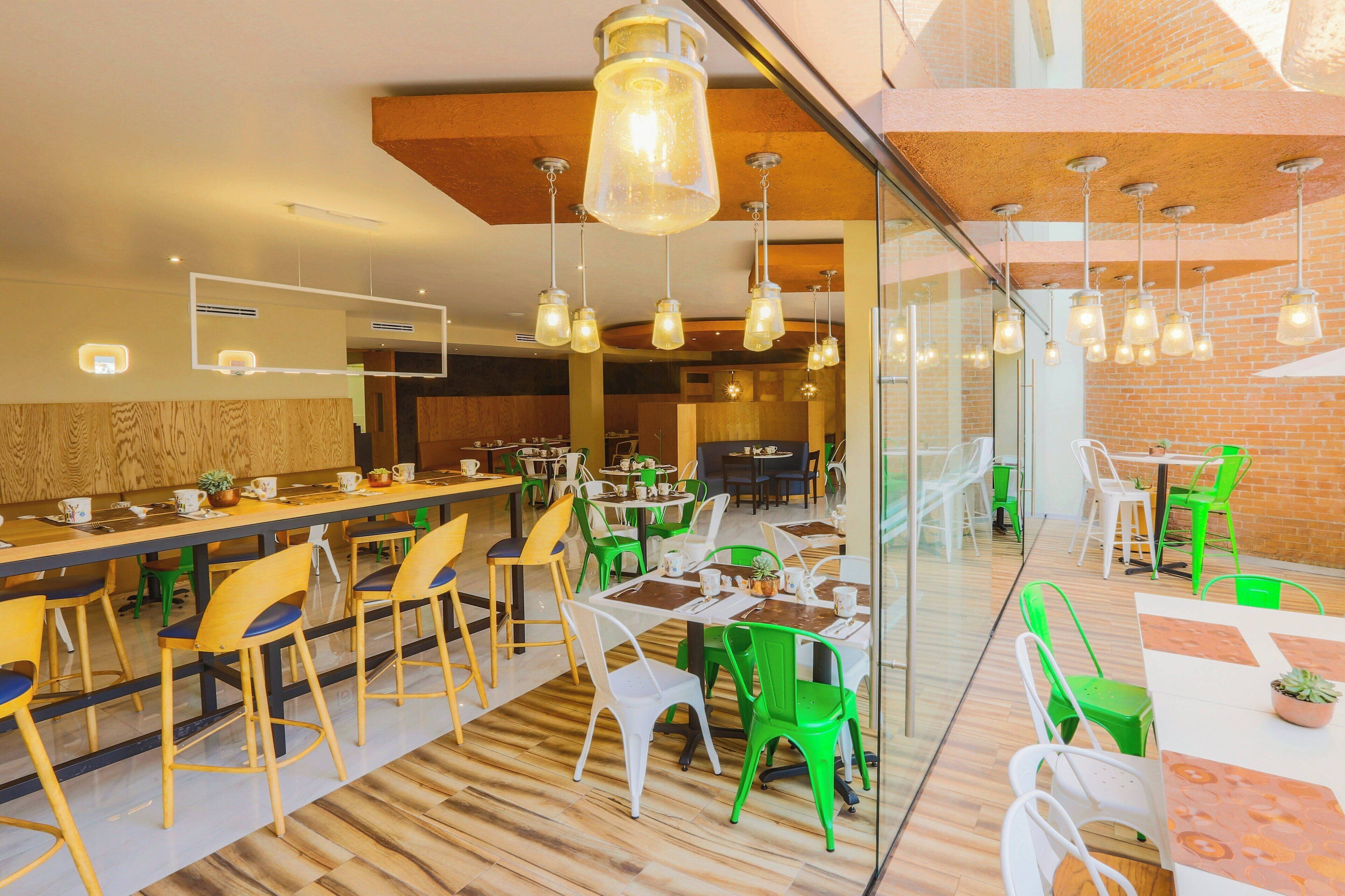 Un espace de restauration moderne avec des tables et des chaises en bois, des chaises vertes et blanches, et des luminaires suspendus.