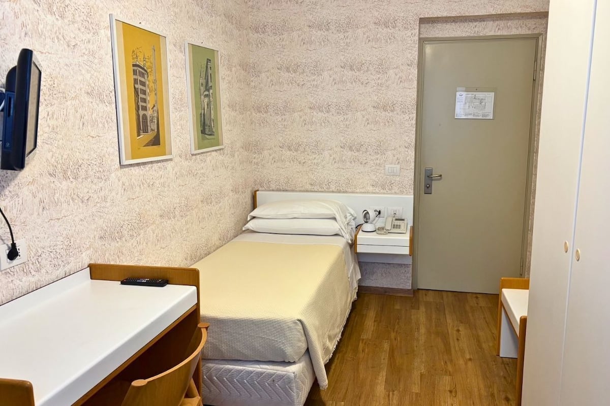 A small, compact room with a bed, bedside table, and a desk with a chair. There is a door with a sign on it, and two framed pictures on the wall.
