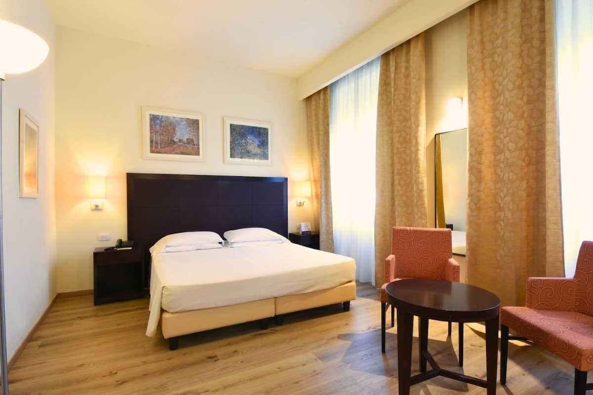 A hotel room with a bed, two bedside tables, a desk, a chair, a round table, and two framed pictures on the wall.