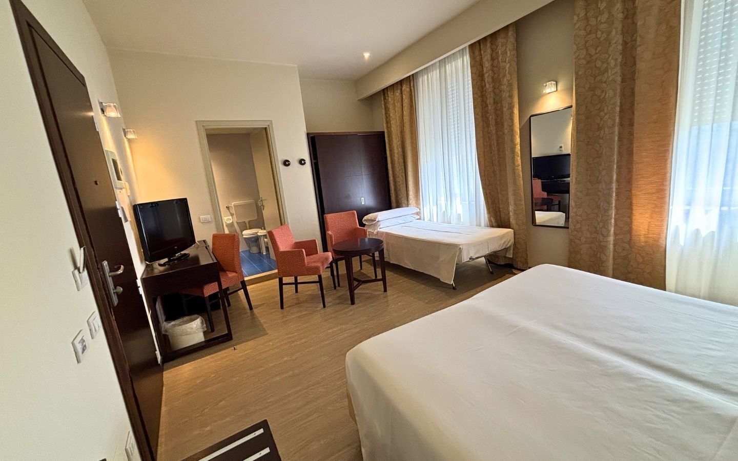 Habitación de hotel con cama, escritorio con silla, televisor y baño visible a través de una puerta abierta.