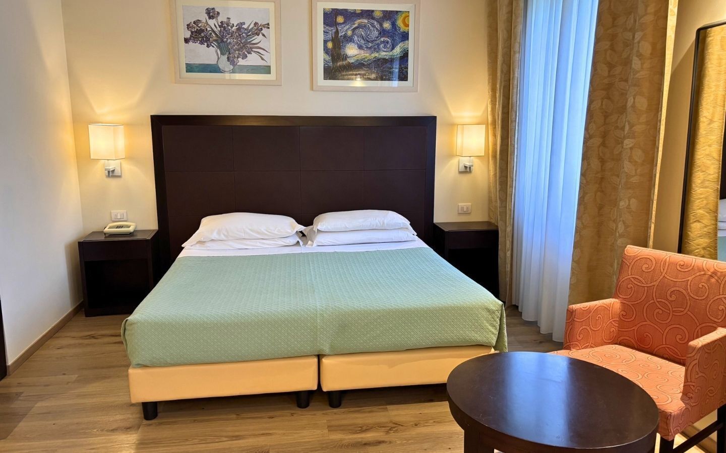 Ein Hotelzimmer mit einem Bett, Nachttischen, einem Sessel, einem runden Tisch und zwei gerahmten Bildern an der Wand.