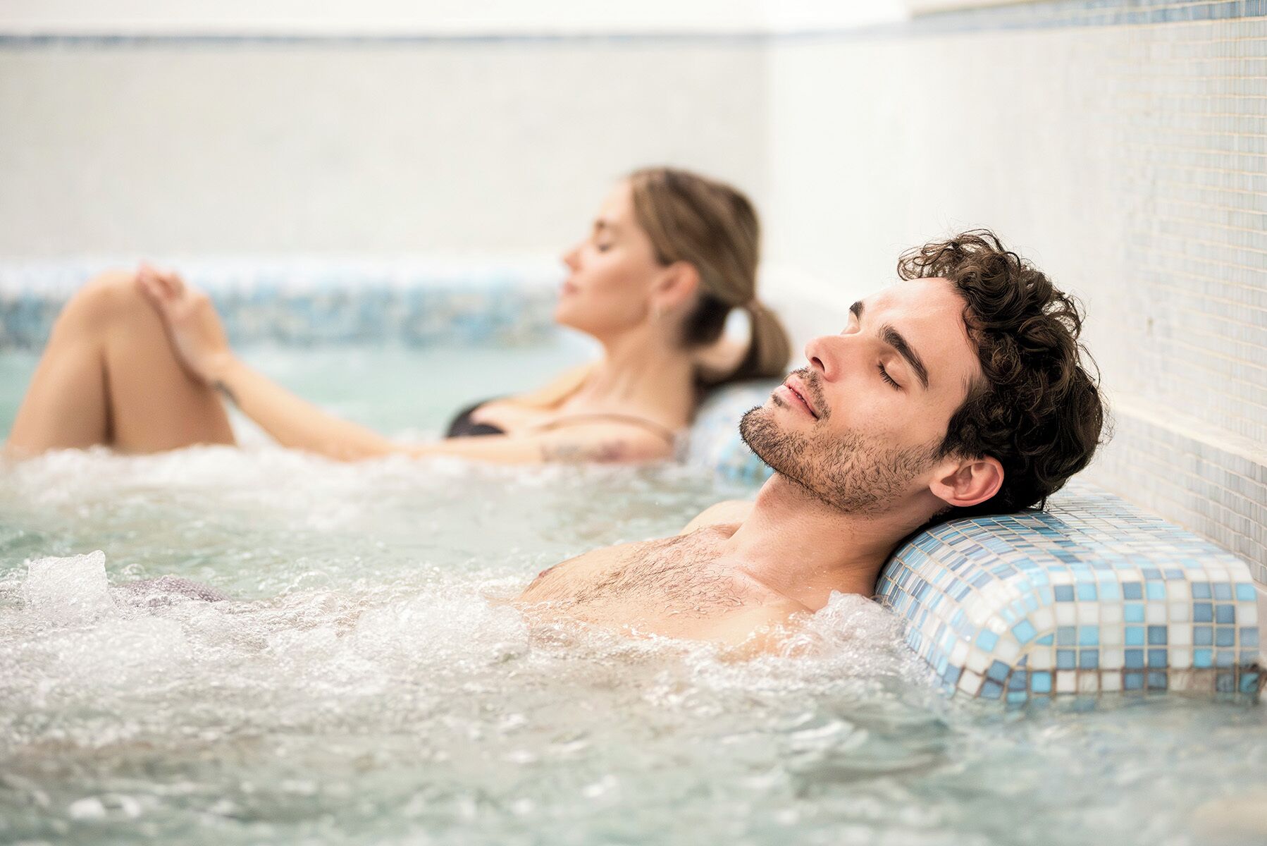 A person relaxing in a hot tub with a tiled wall and a blue mosaic pillow.