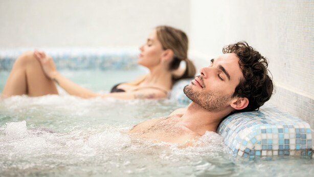 A person relaxing in a hot tub with a tiled wall and a blue mosaic pillow. - Hotel Terme (Montechiarugolo)
