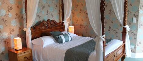 Una cama con dosel sobre cuatro columnas, ropa de cama blanca y una manta verde, con mesitas de noche y lámparas a cada lado.