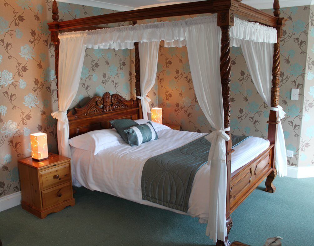 A four-poster canopy bed with a wooden frame and white drapes, flanked by nightstands with lamps, in a room with floral wallpaper.