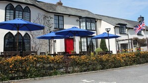 A building with a white facade, black-framed windows, and a red door. There are blue umbrellas and outdoor seating. - Lifton Hall (Lifton)