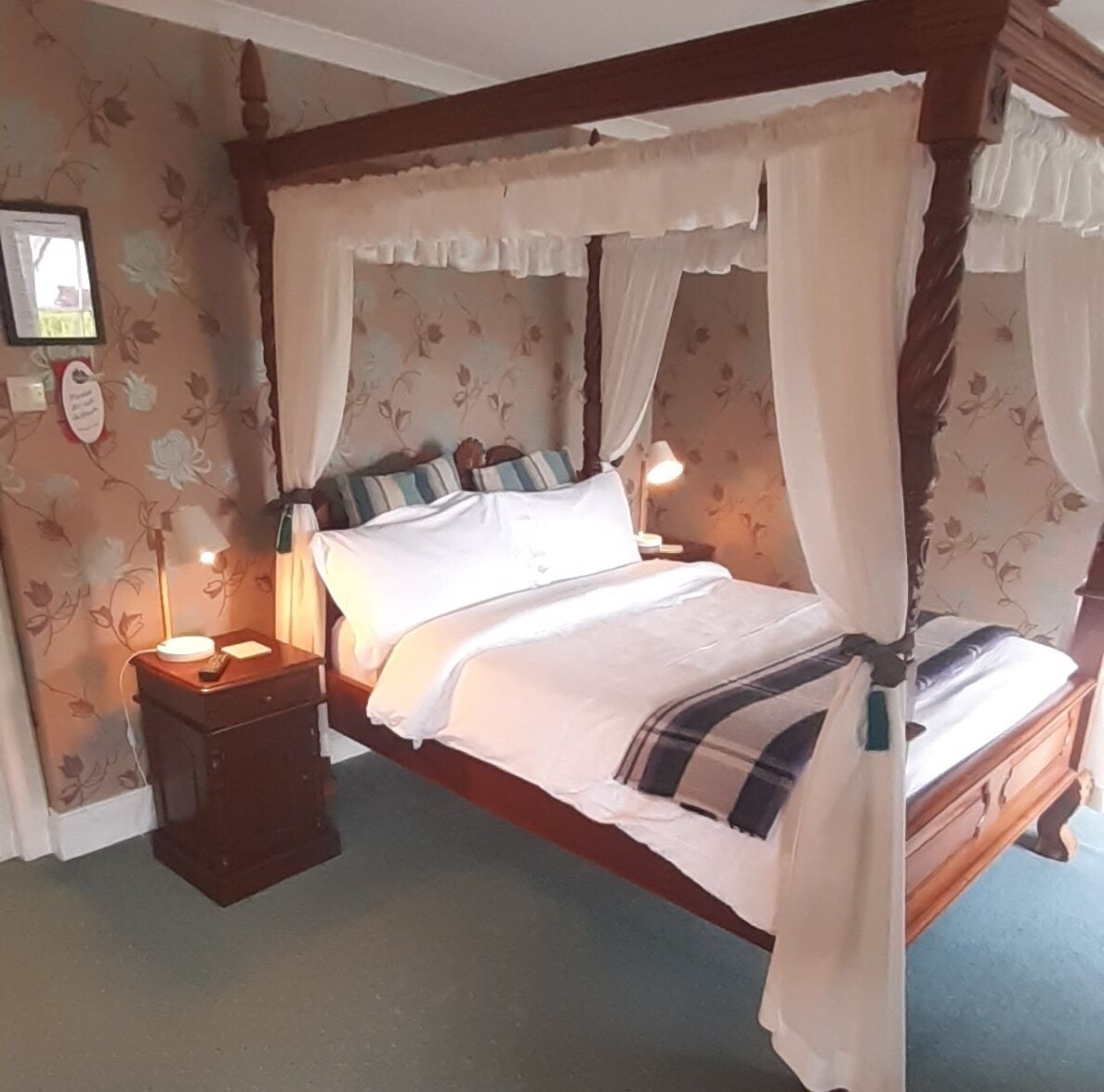 A four-poster canopy bed with white bedding, a nightstand with a lamp, and floral wallpaper.