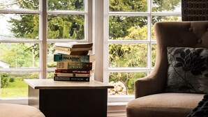 A room with a window, a stack of books on a table, a chair with a floral pillow, and a view of greenery outside. - Lifton Hall (Lifton)