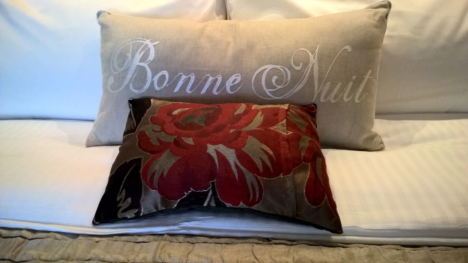Una cama con sábana blanca y dos almohadas, una con diseño floral y la otra con texto bordado.