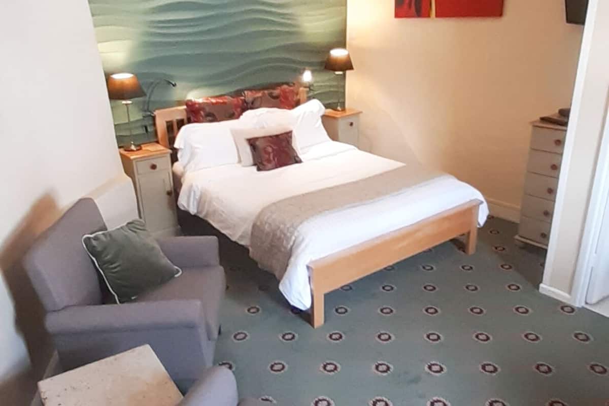 A bedroom with a bed, a chair, a nightstand, a lamp, a painting, and a patterned carpet.
