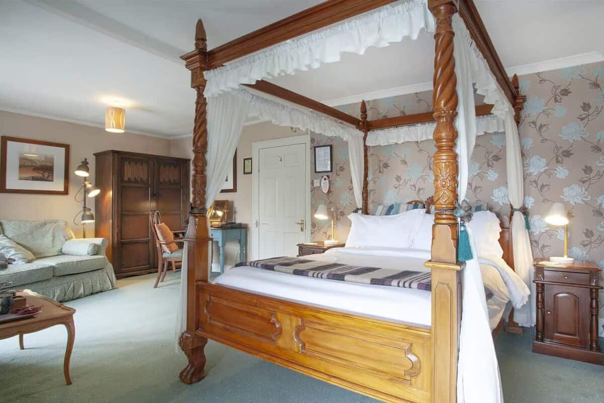A four-poster bed with a canopy, a wooden nightstand, a desk, and a chair in a room with floral wallpaper.