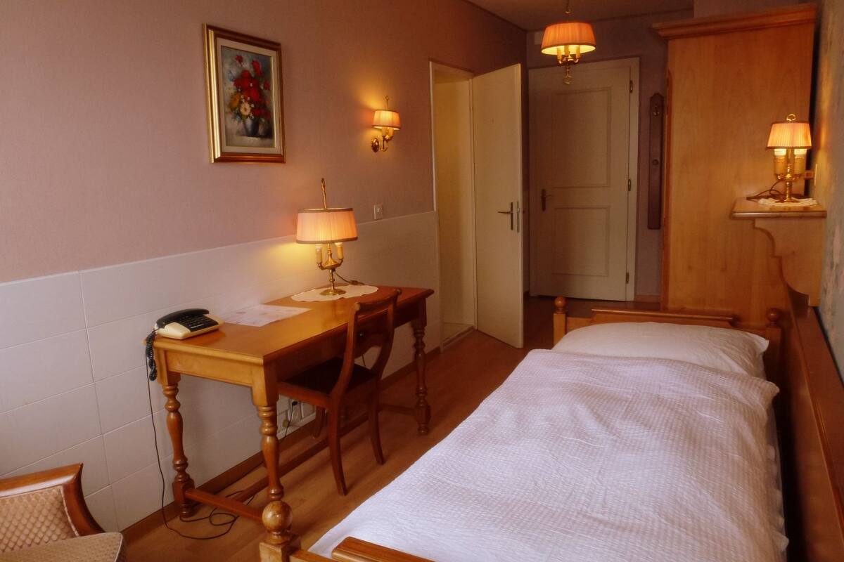A hotel room with a bed, a desk with a lamp, a chair, a telephone, and a door leading to another room.