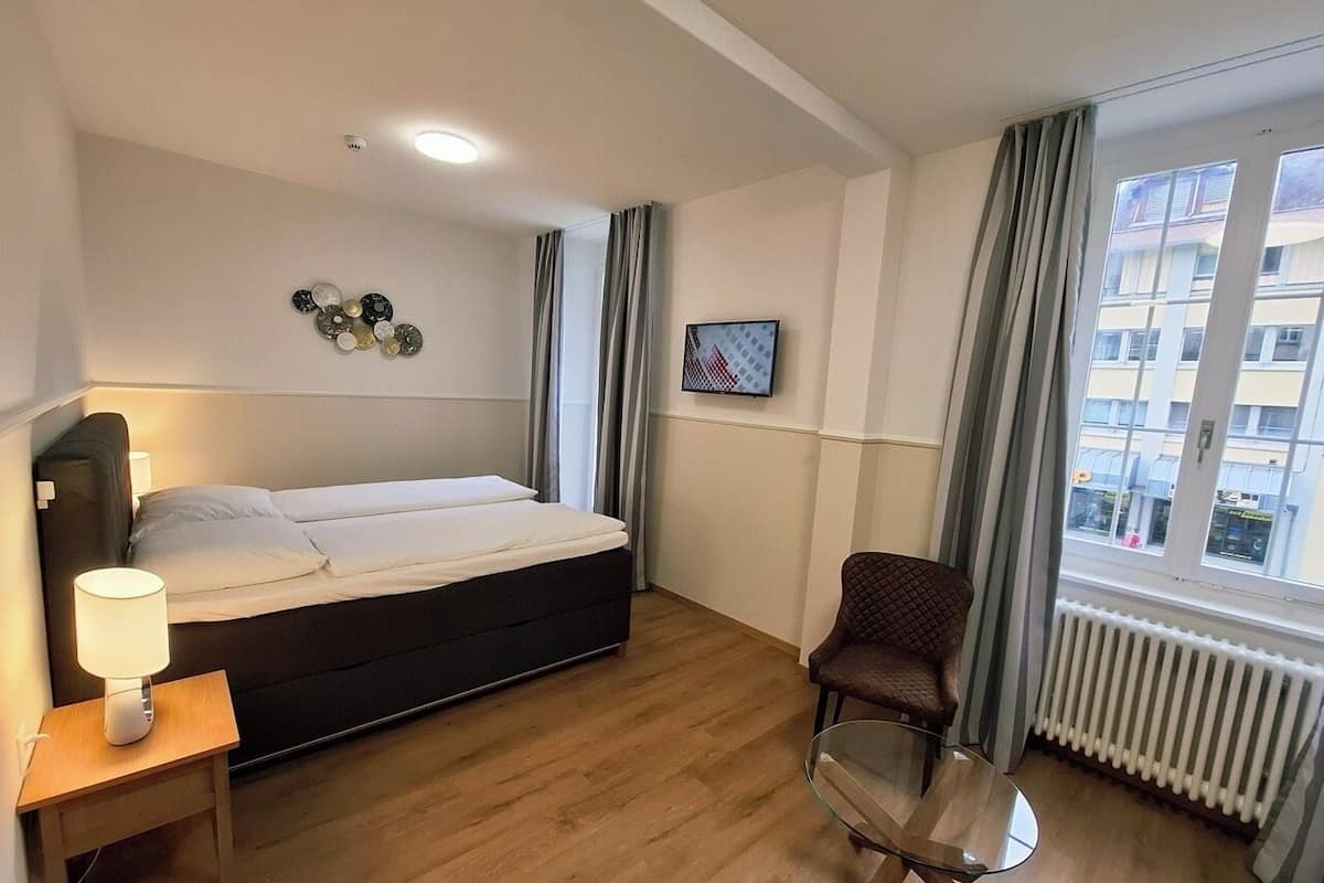 A hotel room with a bed, a chair, a table, a lamp, a picture on the wall, and a window with curtains.