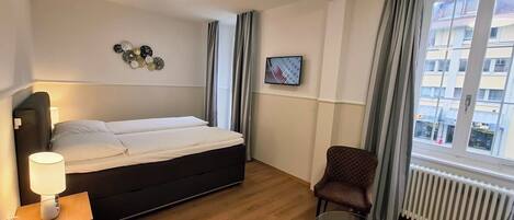 Habitación de hotel con cama, una silla, una mesa, una lámpara, un cuadro en la pared y una ventana con cortinas.