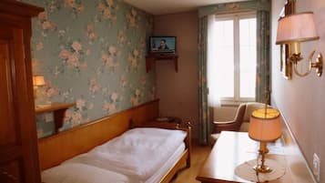 Habitación de hotel con una cama individual, una mesita de noche de madera, un escritorio con lámpara, una silla, un televisor en la pared y una ventana con cortinas.
