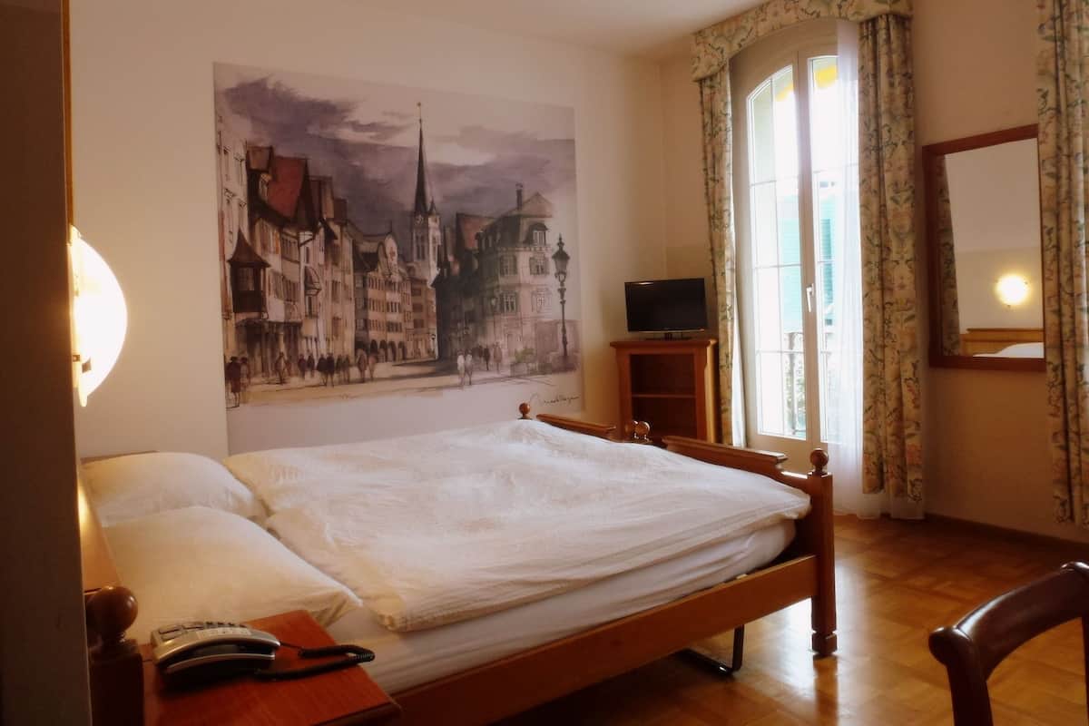 A hotel room with a bed, a television on a wooden stand, a telephone, and a painting of a cityscape on the wall.
