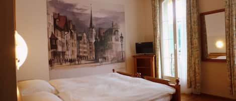 A hotel room with a bed, a television on a wooden stand, a telephone, and a painting of a cityscape on the wall.