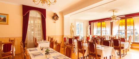 A dining area with multiple tables and chairs, a chandelier, and large windows with curtains.