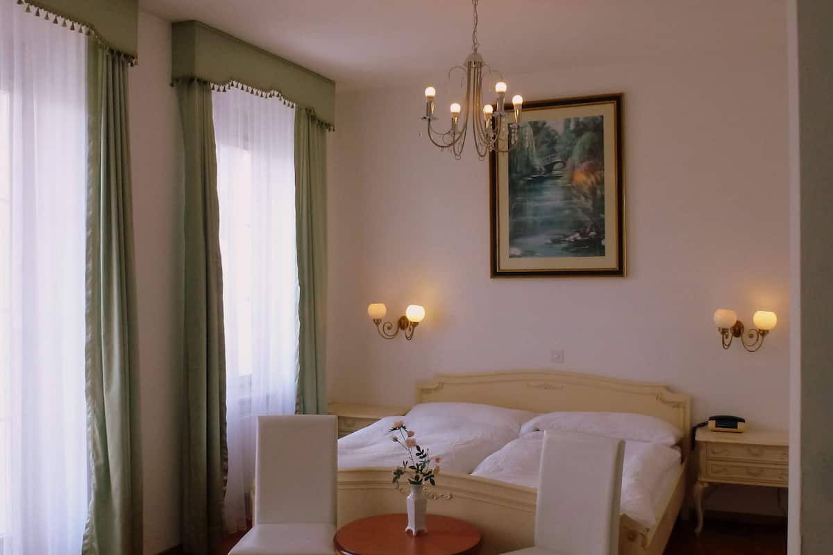 A hotel room with a bed, two bedside tables, a chandelier, a painting, and a table with a vase of flowers.