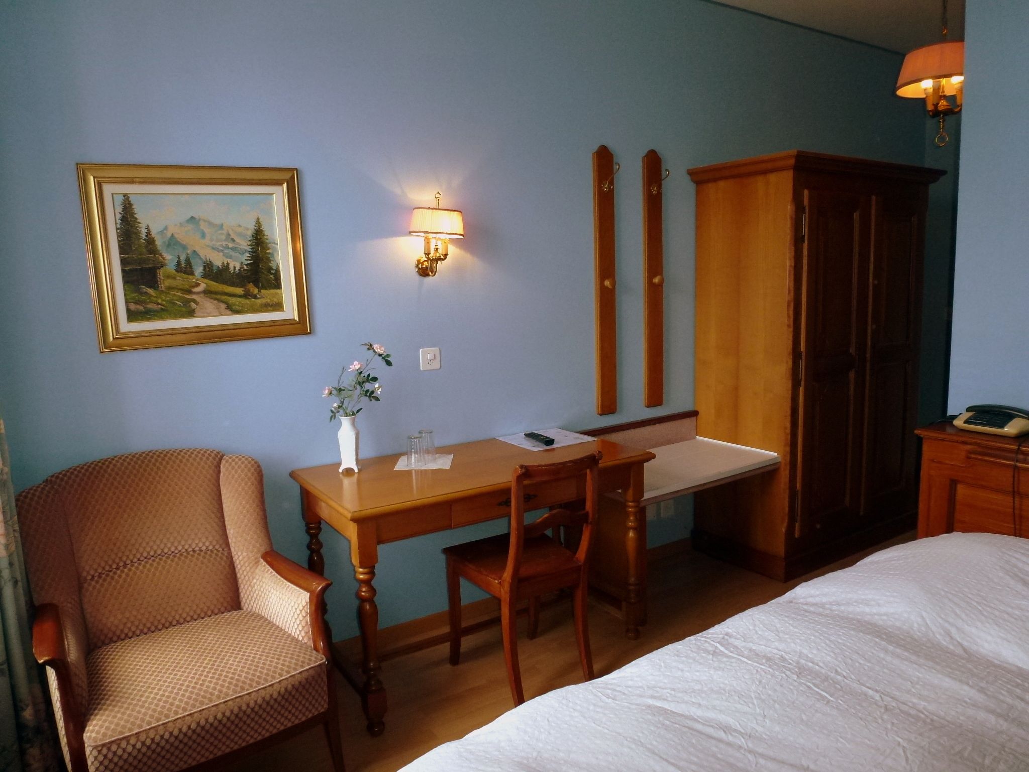 Una camera d'hotel con un letto, una scrivania, una sedia, un armadio, un quadro e un vaso con fiori.