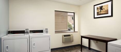 A laundry room with two front-loading washers, a window with blinds, a framed picture on the wall, and a small table.