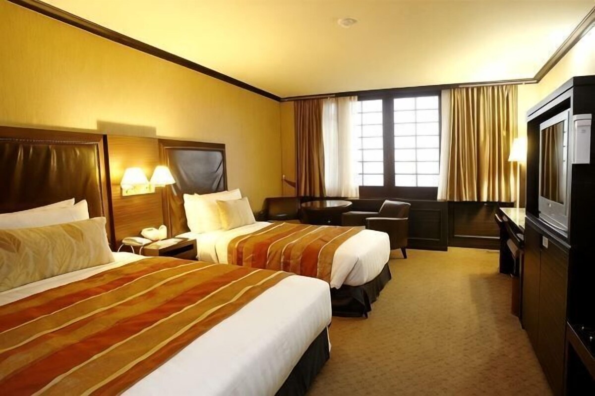 Ein Hotelzimmer mit zwei Betten, einem Schreibtisch, einem Sessel, einem Fernseher und einem Fenster mit Vorhängen.