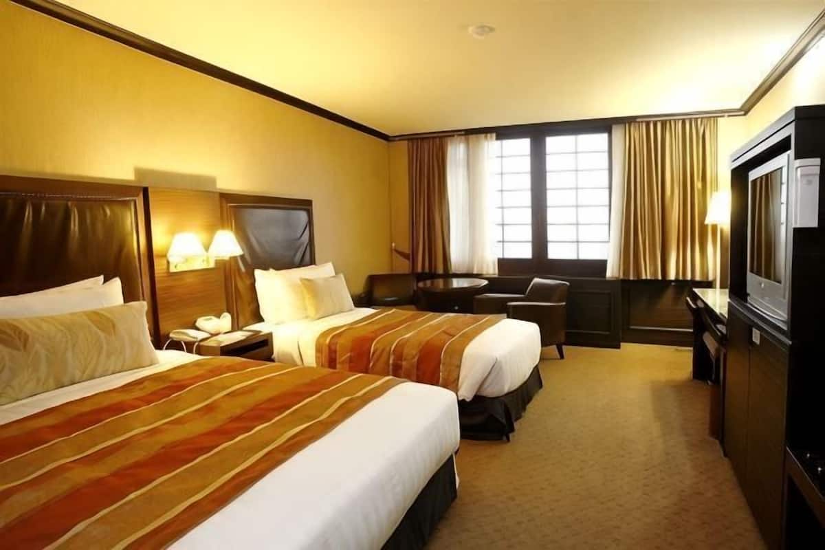 Quarto de hotel com duas camas, uma escrivaninha, uma cadeira, uma televisão e uma janela com cortinas.