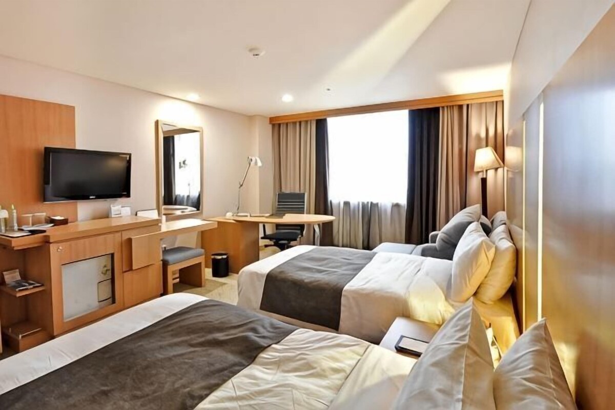 Ein Hotelzimmer mit zwei Betten, einem Schreibtisch, einem Fernseher und einem großen Fenster mit Vorhängen.