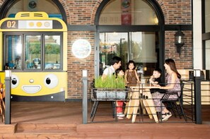 A family dining at an outdoor cafe with a brick building and a yellow bus in the background. - Hotel Miranda (Icheon)