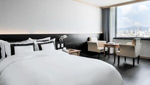 A hotel room with a large bed, a desk, two chairs, a lamp, and a view of a cityscape through a large window. - Hotel Miranda (Icheon)