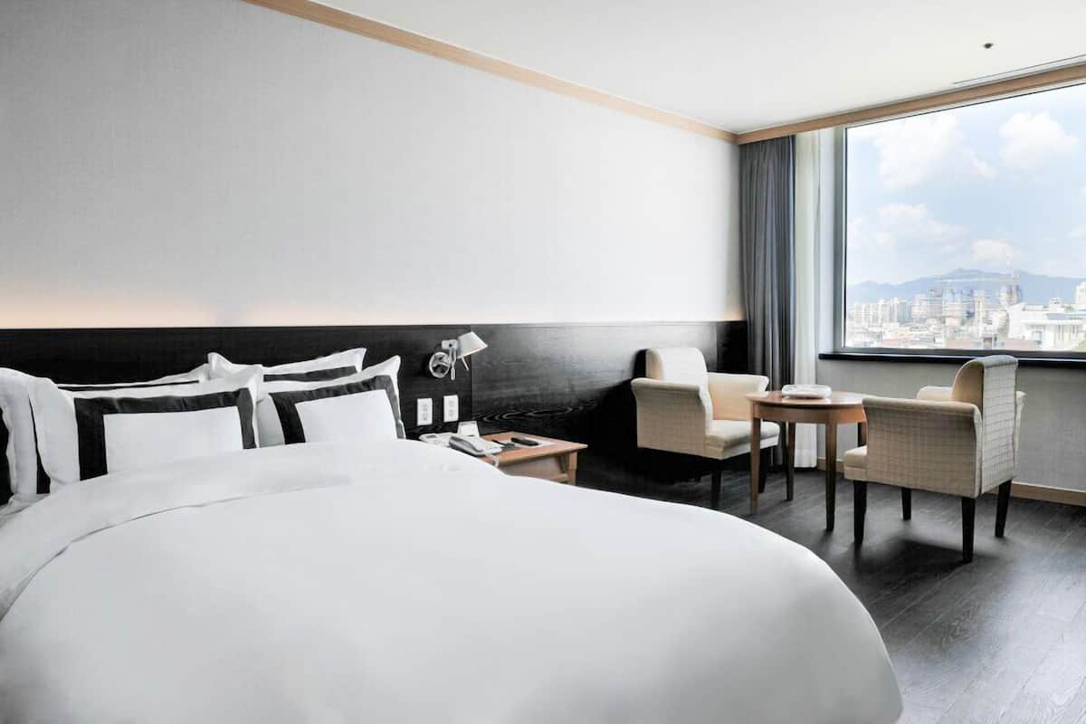 A hotel room with a large bed, a desk, two chairs, a lamp, and a view of a cityscape through a large window.