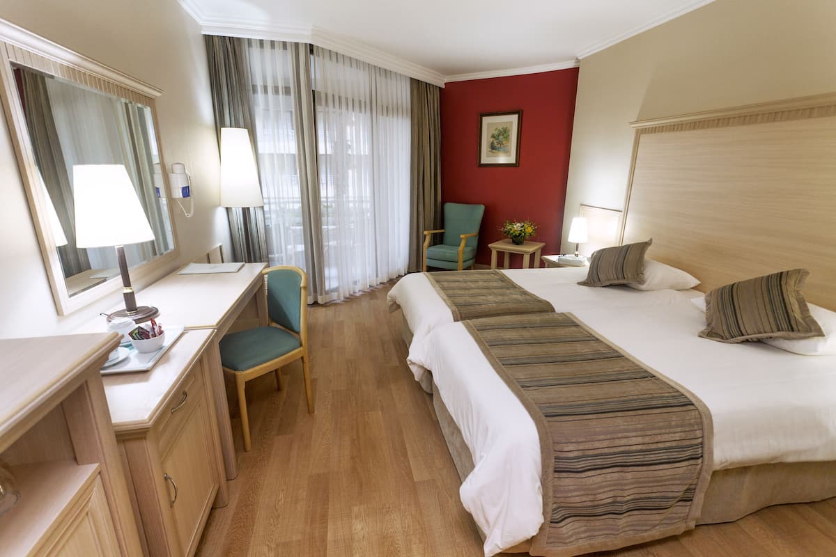 Quarto de hotel com escrivaninha, duas camas, uma cadeira, uma mesinha e janela com cortinas.