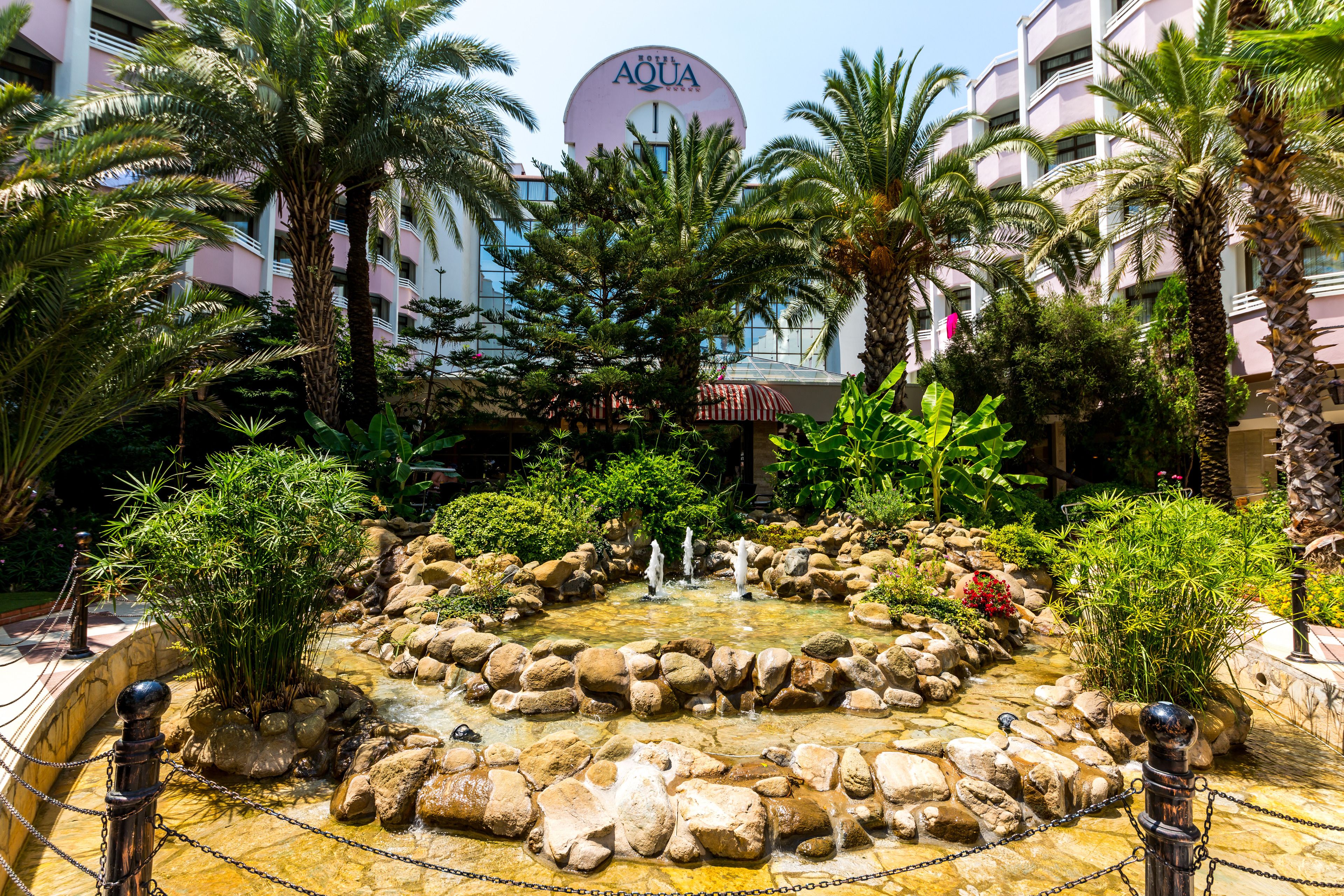 A hotel courtyard with a central water feature surrounded by rocks and palm trees, and a large building with an 'AQUA' sign in the background.
