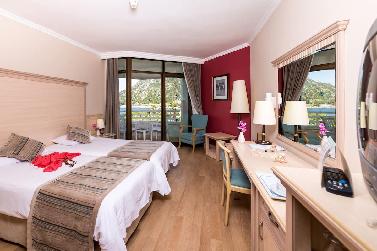 Quarto de hotel com duas camas, uma escrivaninha, uma cadeira, uma mesa pequena, um espelho e vista para o exterior.