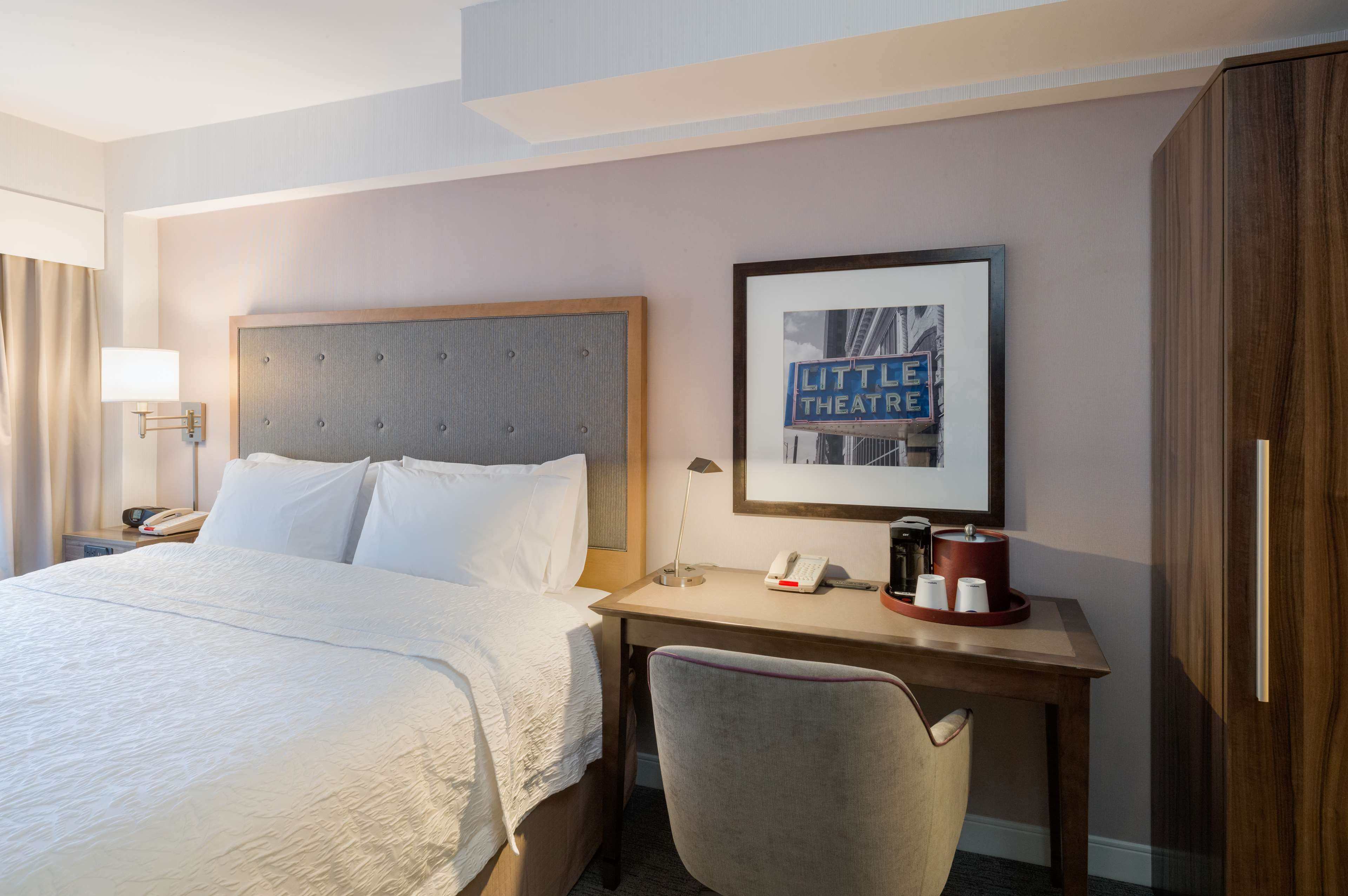 Quarto de hotel com uma cama grande, uma escrivaninha com telefone, um quadro na parede e um armário.