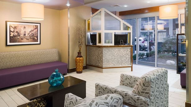 A modern lobby with a purple sofa, patterned armchairs, a coffee table, and a reception desk with a glass partition. - Hampton Inn Madison Square Garden Area Hotel (New York)