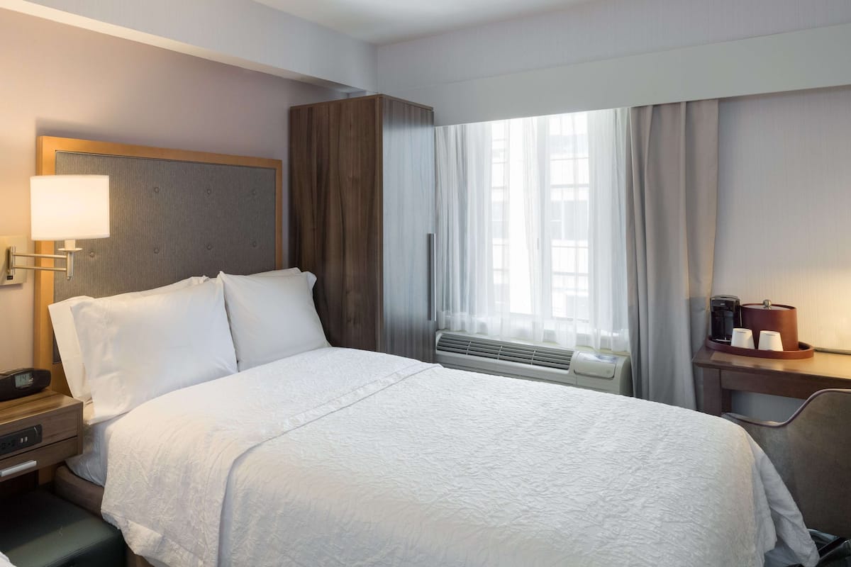 Una habitación de hotel con cama, mesita de noche, lámpara, ventana con cortinas y termostato.