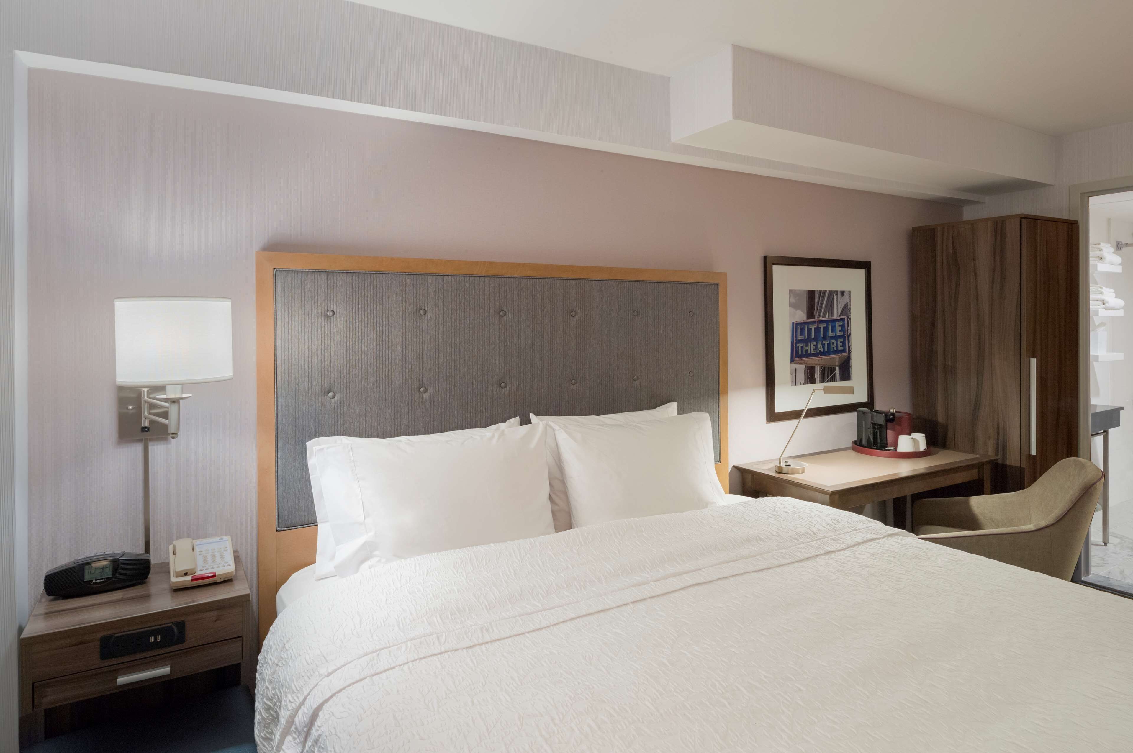 A hotel room with a large bed, a desk with a chair, a nightstand with a lamp, and a wardrobe.