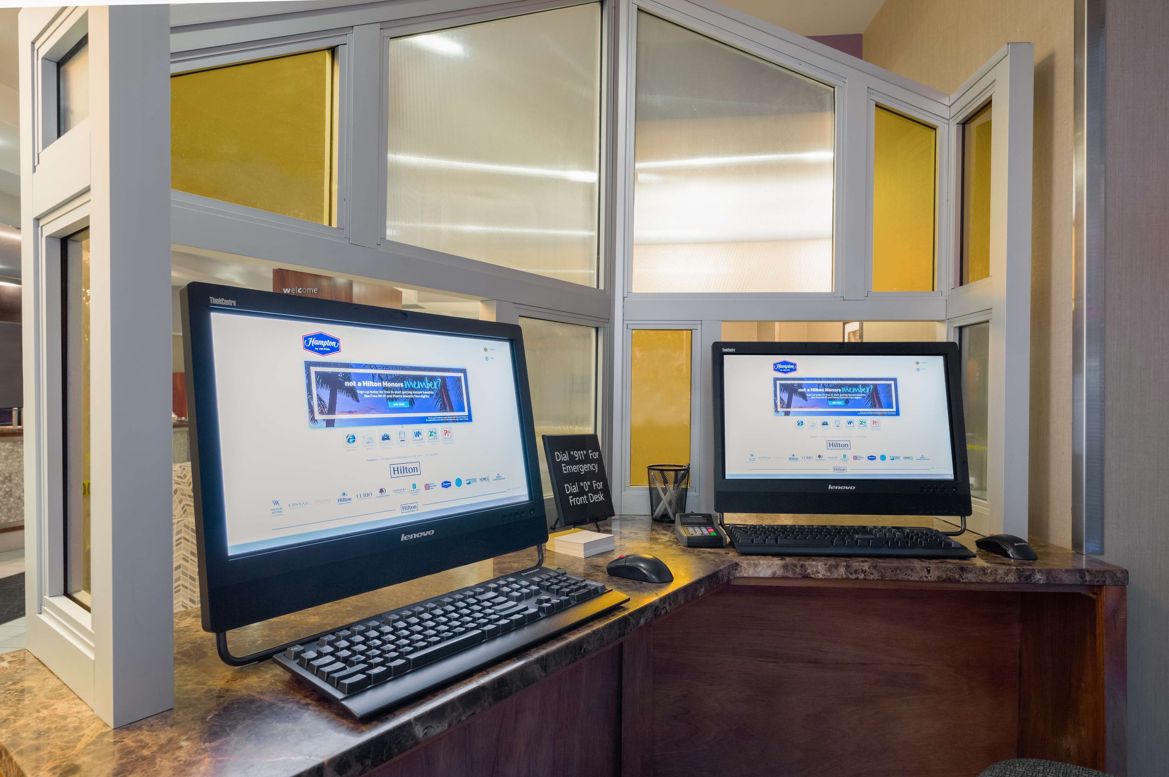 Two computer monitors at a reception desk with a sign indicating a 911 emergency phone.