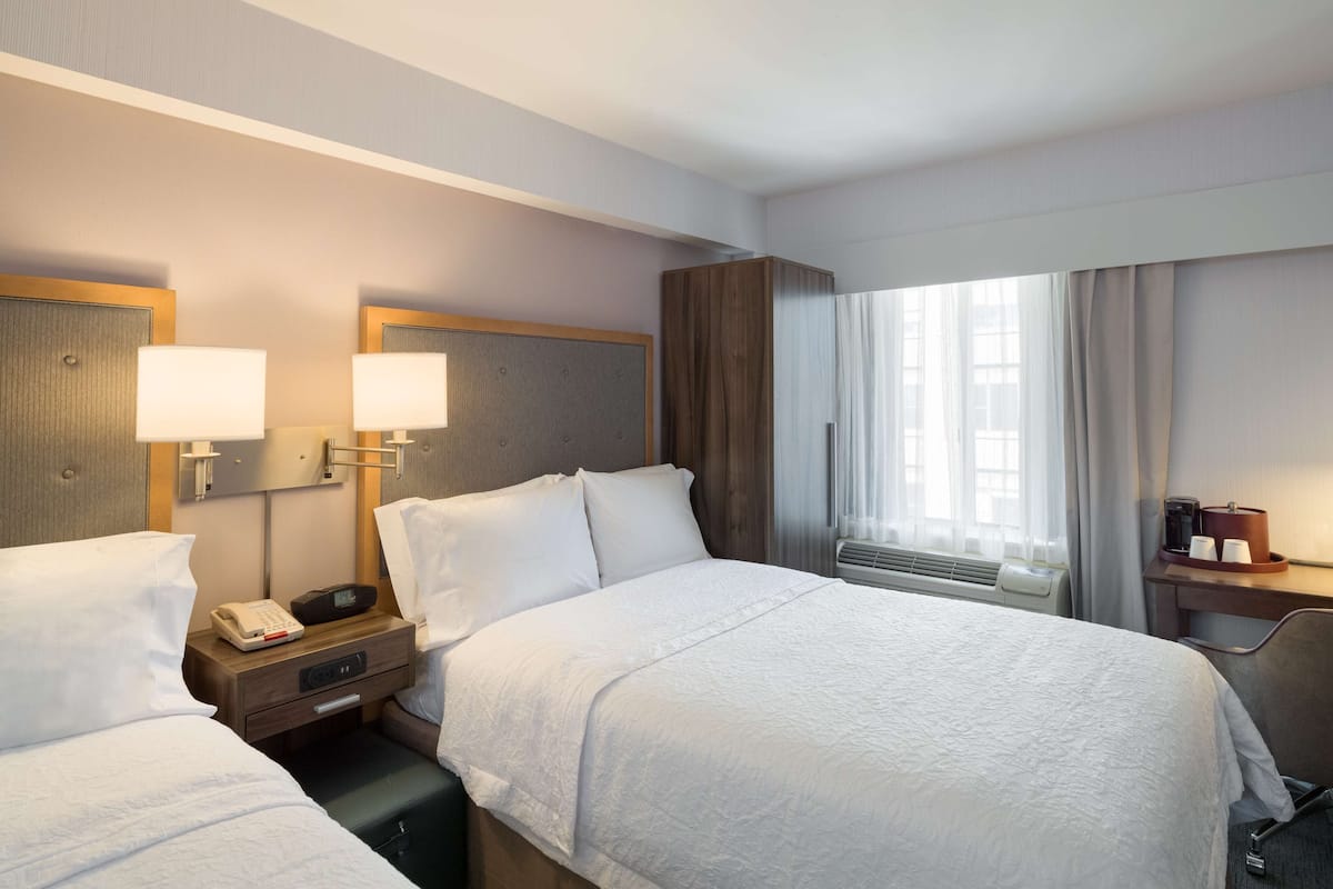 Una habitación de hotel con dos camas, una mesita de noche con un teléfono y una lámpara, una ventana con cortinas y una pequeña mesa con cafetera.